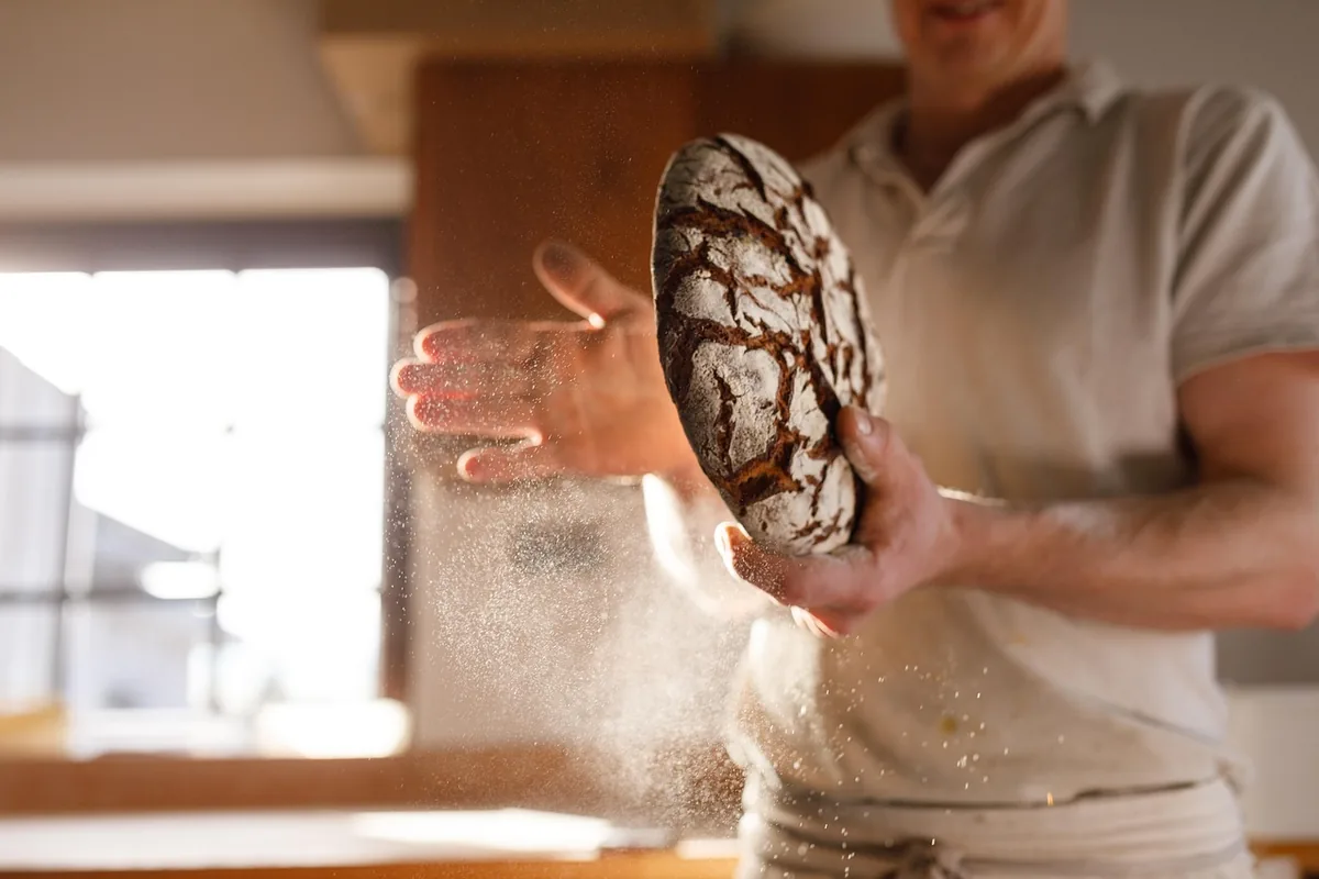 Frieder Kühnast hält ein rundes Roggenbrot mit gerissener Mehlkruste, Mehl staubt im Fensterlicht