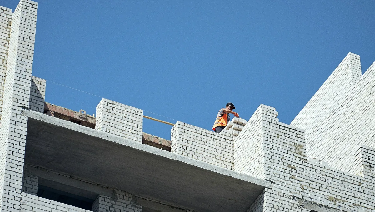 Rohbau aus hellem Kalksandstein unter blauem Himmel, oben auf der Mauer setzt ein Maurer in Warnweste die nächsten Steine.
