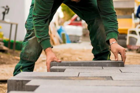Ein Pflasterer in grüner Arbeitskleidung verlegt Betonsteinpflaster.
