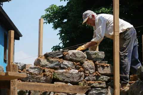 Ein älterer Maurer setzt eine Natursteinmauer mit Mörtel neu auf, entlang einer gespannten Schnur.