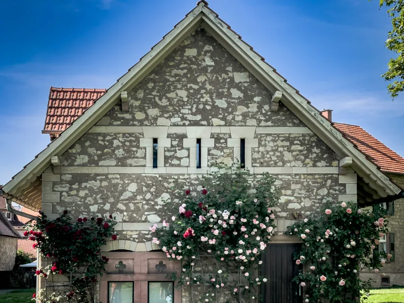 Altes Haus mit Bruchsteinfassade unter blauem Himmel, davor blühende Kletterrosen.