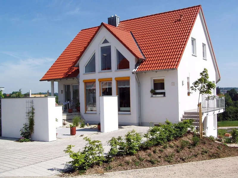 Fertiges, weiß verputztes Einfamilienhaus mit rotem Ziegeldach, davor eine frisch gepflasterte Hoffläche und ein neu bepflanzter Hang.