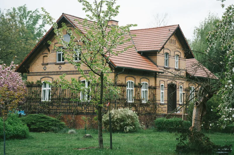 Gelbes Backsteinhaus mit Giebeln und rotem Ziegeldach hinter einem grünen Garten mit Obstbaum.
