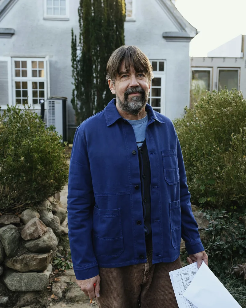 Martin Riedholm in blauer Arbeitsjacke vor einem weißen Haus, in der Hand ein Plan.