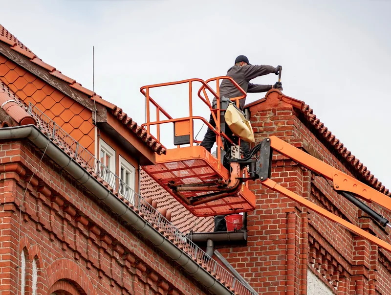 Handwerker auf einer Arbeitsbühne am Giebel eines Backsteinhauses mit roten Ziegeln, daneben Dachrinne und Fallrohr.