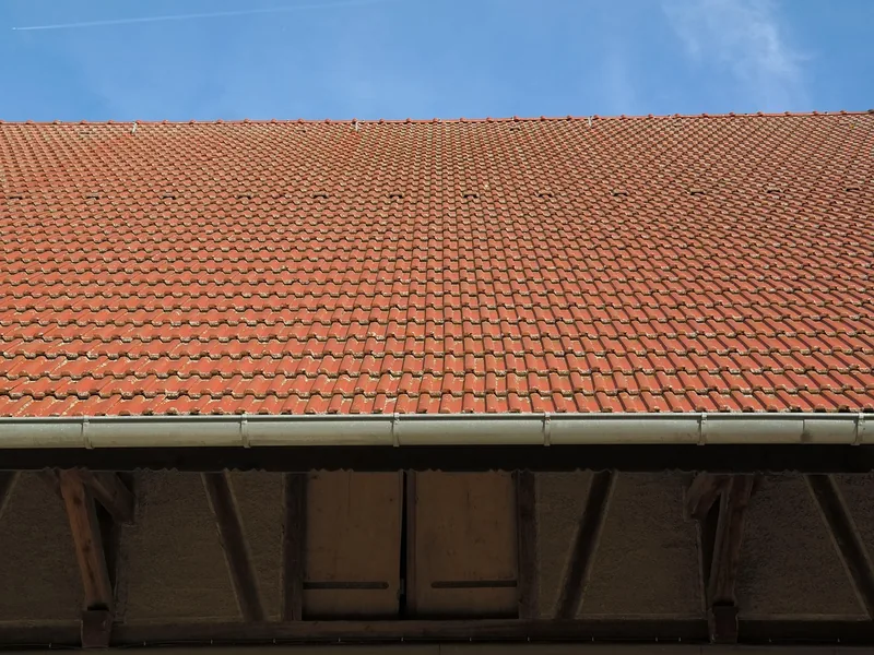 Langes Ziegeldach mit verzinkter Rinne an der Traufe, darunter dunkle Holzbalken. Blauer Himmel.