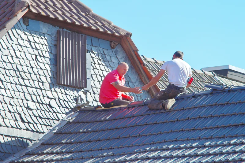 Zwei Dachdecker sitzen auf einem Dach neben einer Gaube und reichen sich etwas an. Blauer Himmel.
