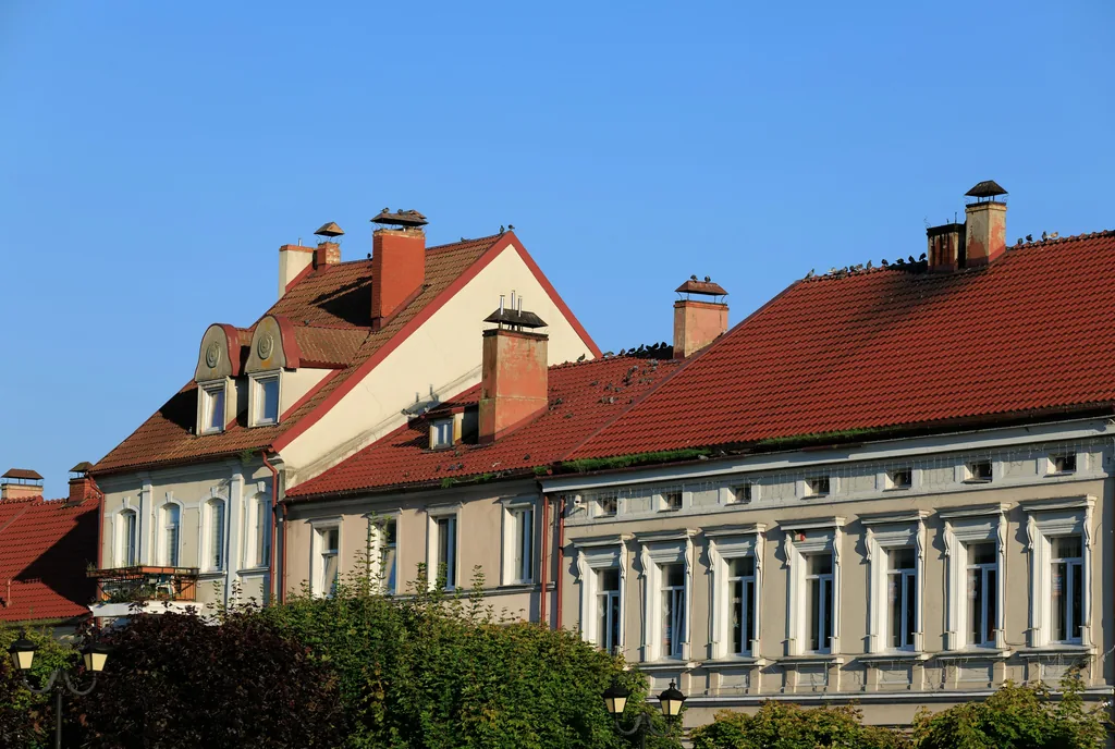Zwei historische Stadthäuser mit hellen Fassaden, roten Ziegeldächern, Gauben und Schornsteinen, davor Bäume und Laternen. Blauer Himmel.