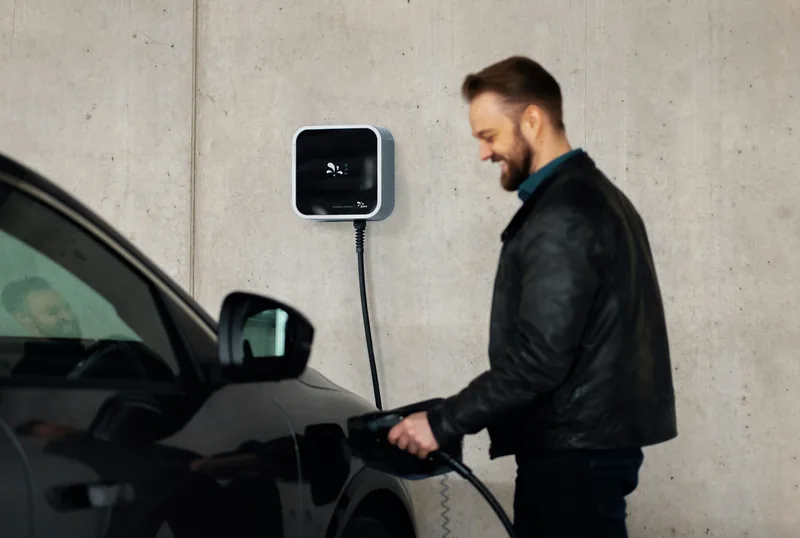 Wallbox an der Wand, ein Mann steckt lächelnd das Ladekabel in sein Auto.