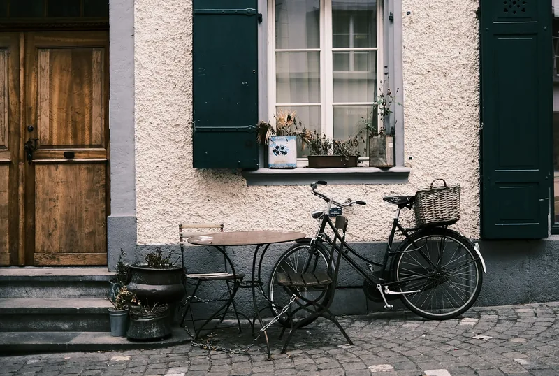 Ein Fahrrad mit Korb vor einem hellen Altstadthaus mit Holztür, dunklen Fensterläden und Kopfsteinpflaster.