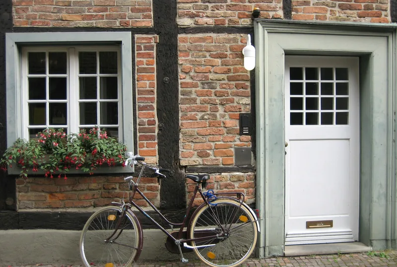 Ein Fahrrad lehnt an der Backsteinfassade neben der weißen Eingangstür, unter dem Fenster blüht ein Blumenkasten.
