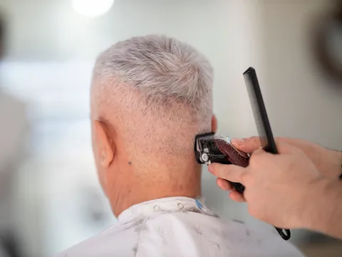 Ein älterer Herr mit grauem Kurzhaarschnitt von hinten, im Nacken wird mit Kamm und Maschine nachgeschnitten.
