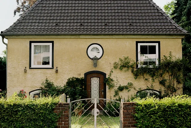 Gelb verputztes Haus mit dunklem Ziegeldach, Hecke und schmiedeeisernem Gartentor