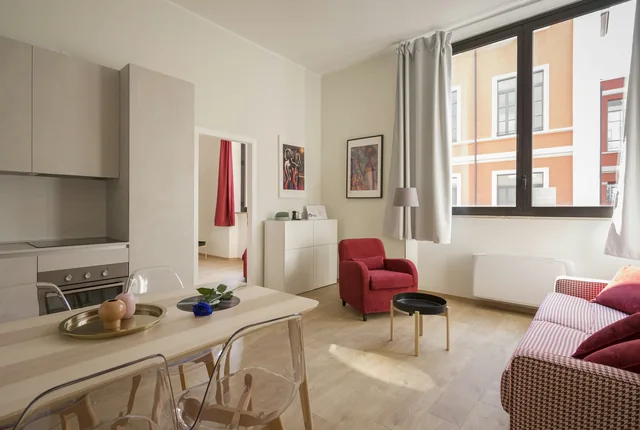 Helles Wohnzimmer mit Essplatz, rotem Sessel und hohem Fenster, gegenüber Altbaufassaden