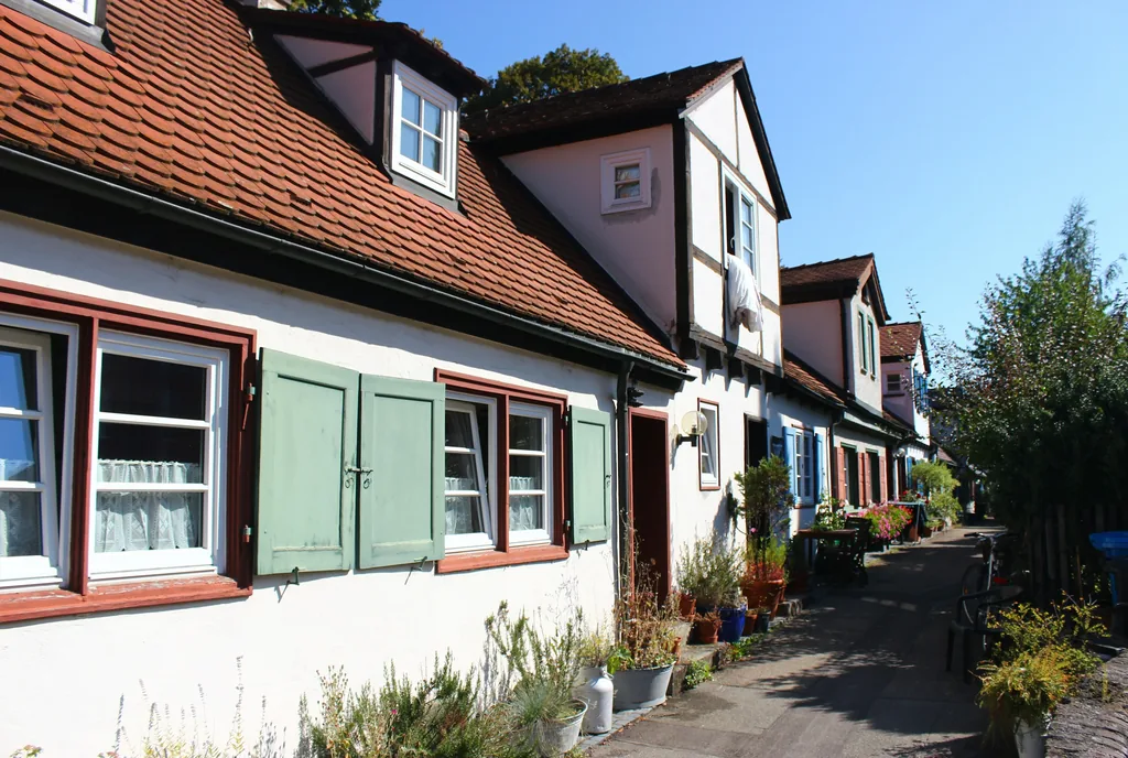 Kleine Siedlungshäuser mit Gauben, grünen Fensterläden und Blumentöpfen an einem schmalen Weg