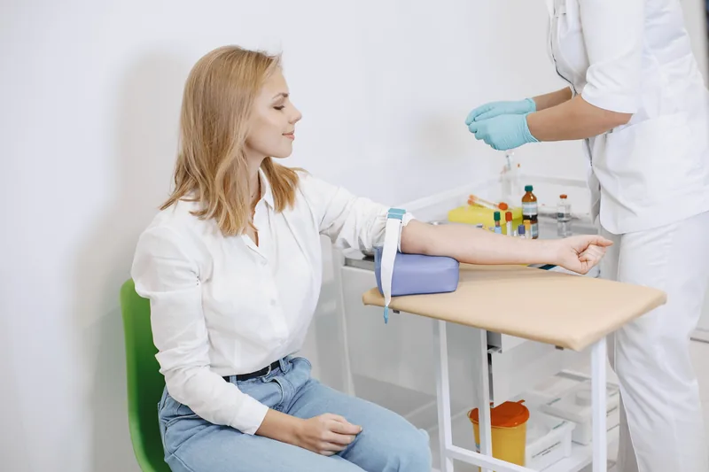 Eine Patientin sitzt mit Stauband am Arm, eine Mitarbeiterin in Weiß bereitet die Blutabnahme vor