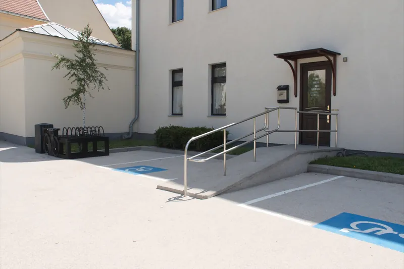 Hof hinter der Praxis: Rampe mit Handlauf zur Hintertür, breiter Parkplatz mit Rollstuhlzeichen, Fahrradständer und eine junge Birke