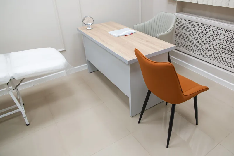 Helles Sprechzimmer mit Schreibtisch, Untersuchungsliege und einem orangefarbenen Stuhl