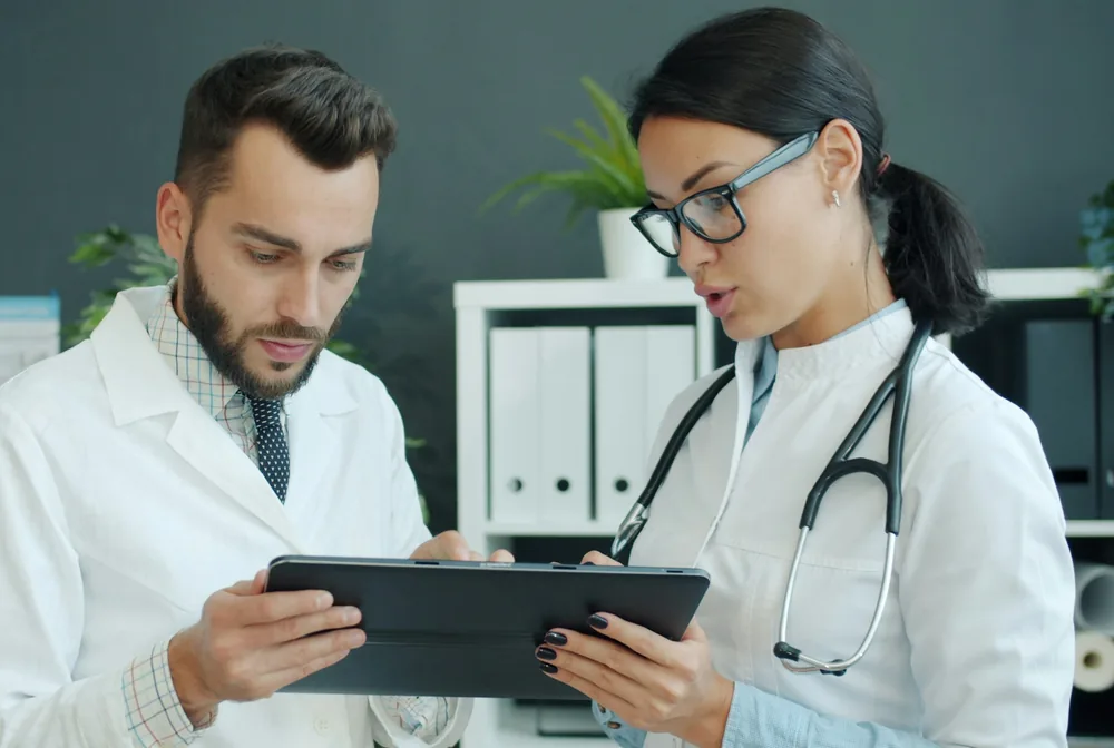 Eine Ärztin und ein Arzt im weißen Kittel schauen im Sprechzimmer gemeinsam auf ein Tablet